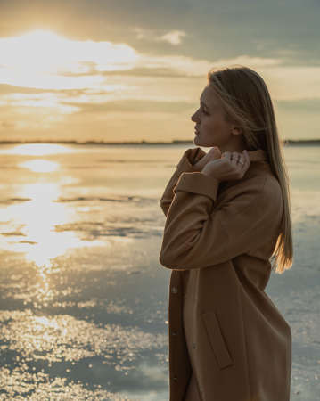 Beautiful young blonde girl with long hair walks along the lake shore and looks at the sunset. Autumn walk. The girl in the coatの写真素材