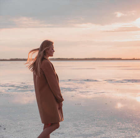 Beautiful young blonde girl with long hair walks along the lake shore and looks at the sunset. Autumn walk. The girl in the coatの写真素材