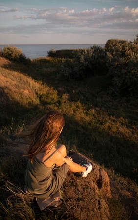 Young girl in a tank top, sitting on a hill. Beautiful views and sunrise or sunset. Girl with long hair. The view from the backの写真素材