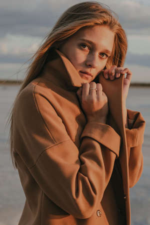 Beautiful young blonde girl with long hair walks along the lake shore and looks at the sunset. Autumn walk. The girl in the coatの写真素材