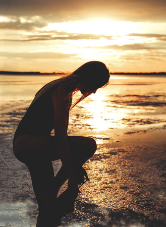 Beautiful young blonde girl with long hair walks along the lake shore and looks at the sunset....
