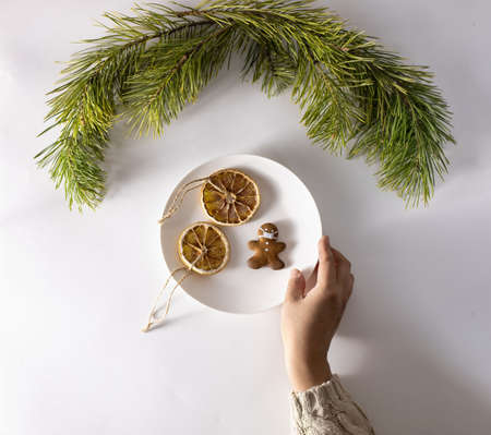 Womans hand in a knitted sweater holds a plate of Christmas sweets. Cookies in the form of a man in a mask and dried orange slices with cinnamon. Pine branches on a white backgroundの写真素材