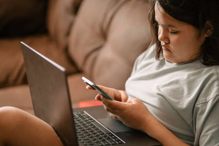 Chubby Asian teen girl is talking on the phone at home, sitting on the sofaの写真素材