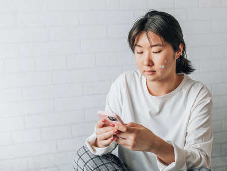 girl is texting on a smartphone while sitting on a bed at home. copy space. teenage girl with problematic skin, with a therapeutic cream applied.の写真素材