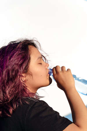 Asian girl with purple drinks water from a plastic bottle on a white isolated background. Generation Z. The concept of thirstの写真素材