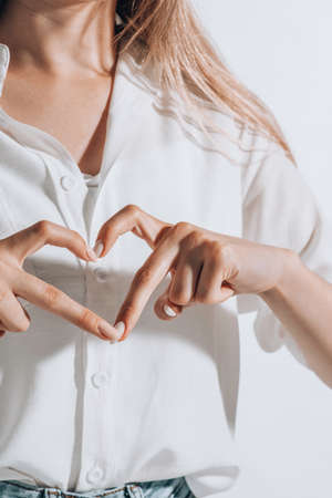 romantic young woman in shirt making a heart gesture with her fingers in front of her chest showing her love and affectionの写真素材