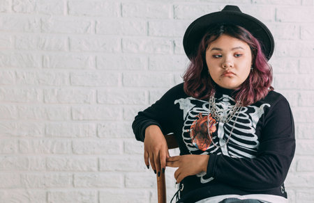 Young Asian teen in costume witch sitting on a chair in front of a white brick wall, background of Halloween.の写真素材