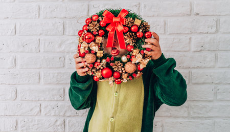 Little boy with a Christmas wreath grimaces, shows his tongue against a white brick wallの写真素材