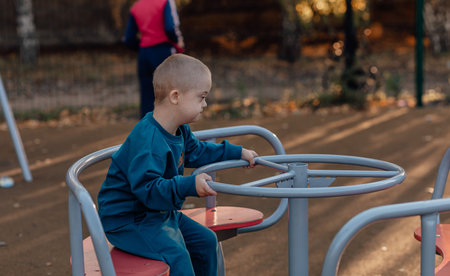 Cute Little boy with down syndrome in a funny hat walks in the playground, spinning on a carouselの写真素材