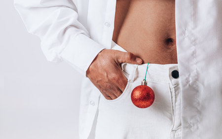Male torso with abs and a red Christmas ball. A man in a white shirt and trousers on a white backgroundの写真素材