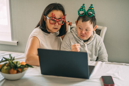 girl helps an elderly woman with Down syndrome in funny Christmas glasses to make a video call from laptop, congratulationsの写真素材