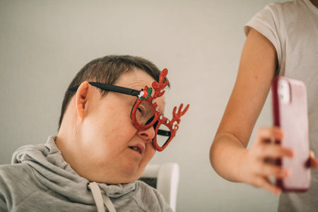 girl helps sleepy elderly woman with Down syndrome in funny Christmas glasses to make a video call from her phone, congratulationsの写真素材