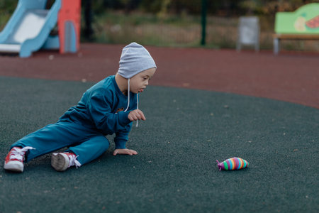 boy with down syndrome is sitting on a rubber covering on the playground, reaching for a toyの写真素材