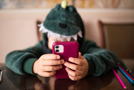 boy who looks into the phone sitting in a dinosaur costume at the table. Unrecognizable faceの写真素材