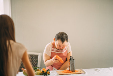 Lifestyle, education. An elderly woman with Down syndrome rubs carrots on a grater with an assistantの写真素材