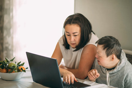 elderly woman with Down syndrome Sitting With Home Tutor Using Laptop For Lesson At Homeの写真素材