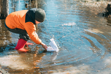 Springtime Sailor : Amid the budding trees, a young boy leans into the experience of launching a paper boat, symbolizing new beginnings and simple joysの写真素材