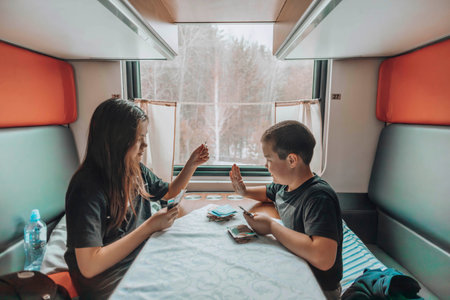 Sibling Bonding Time : In the cozy train compartment, two young individuals engage in a playful card game, surrounded by the serene winter landscape visible through the windowの写真素材