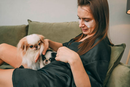 woman in black silk robe grooming fluffy dog on sofa, relaxed morning routine, concept of pet care and leisure timeの写真素材