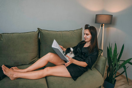 woman with eye patches relaxing on couch, reads book, and petting dog in cozy living room, concept of self-care and pet careの写真素材