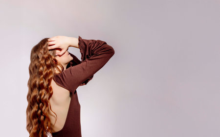 young woman with long wavy red hair touching her head with hand in stylish brown dress with open back on light background, beauty salon, haircare, fashion industry, web bannerの写真素材