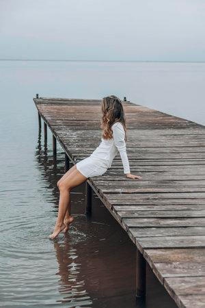 unrecognizable woman in a white dress, sitting on the edge of wooden pier, dipping her feet into calm water with a light splash, gazing at horizon under soft cloudy sky, relaxation, travel, verticalの写真素材
