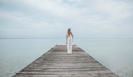 Asian woman standing barefoot on a wooden pier, gazing into the distance over a calm sea horizon, dressed in a white outfit under a pale cloudy sky, mindfulness, wellness retreat, travel blogの写真素材