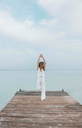 Asian woman stands barefoot with hands up on wooden pier, gazing into distance over calm sea horizon, in a white outfit under a pale cloudy sky, mindfulness, wellness retreat, travel blogの写真素材