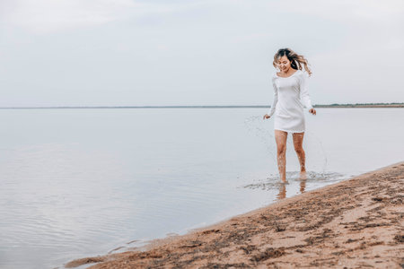 Asian woman in white dress splashing in shallow water at beach, smile, looks down, calm lake with soft horizon under cloudy sky, summer lifestyle, travel, wellness retreats, motion blurの写真素材
