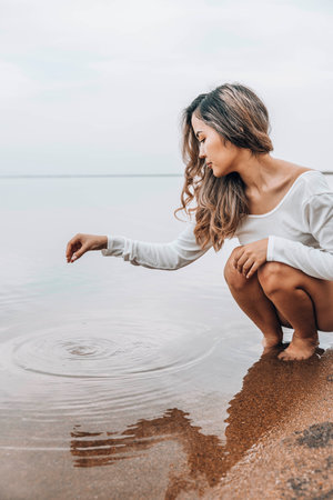 Asian woman squatting barefoot at lake edge creating water ripples with fingers, gazing at water with serene expression, calm horizon under cloudy sky, mindfulness, wellness, nature retreatの写真素材