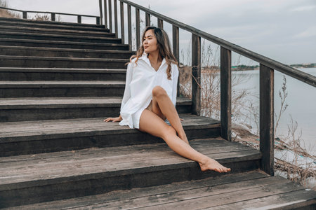 Asian woman sitting barefoot on wooden staircase near lake, gazing into the distance with a relaxed and confident expression, wearing white shirt dress, concept of travel, lifestyle, selfcareの写真素材