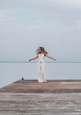 woman dancing barefoot on a wooden pier, long hair flowing as she twirls freely under a pale sky, surrounded by calm water, joyful expression in motion, freedom, wellness, motion blurの写真素材