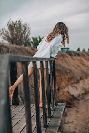 unrecognizable woman in white shirt standing barefoot on wooden platform with one foot raised, natural cliff landscape, back view, solitude, personal reflection, nature retreatの写真素材