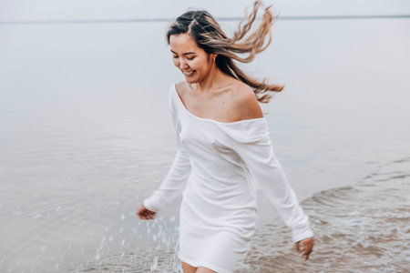 Asian woman in white dress splashing in shallow water at beach, smile, looks down, calm lake with soft horizon under cloudy sky, summer lifestyle, travel, wellness retreats, motion blur, close upの写真素材