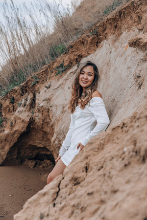 young Asian woman on beige stone wall, smiling gently in white off-shoulder top with relaxed expression, beauty, natural skincare, feminine lifestyleの写真素材