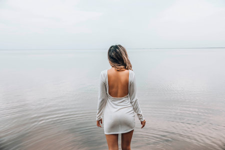 unrecognizable woman standing with back exposed in open white dress near calm lake, facing the water with relaxed posture and serene mood, concept of wellness, spa, fashion editorialの写真素材