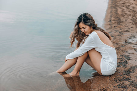 young Asian woman in white dress sitting on sandy lake shore gazing at water with thoughtful expression creating calm serene atmosphere concept of wellness mental health lifestyle blogの写真素材