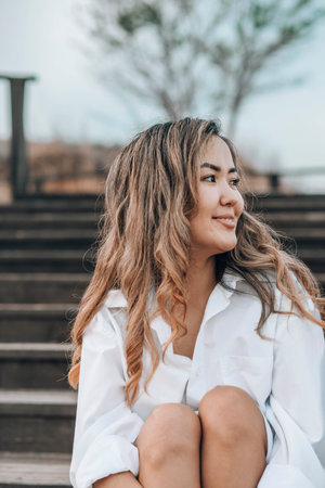 Asian young woman with wavy hair on wooden stairs in white shirt, with hand in hair, peaceful emotional moment outdoors, mindfulness, feminine wellness, emotional balanceの写真素材