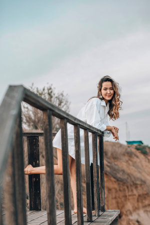 Asian woman in white shirt standing barefoot on wooden platform with one foot raised, natural cliff landscape, back view, solitude, personal reflection, nature retreatの写真素材