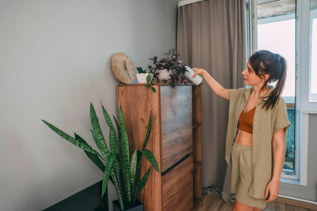 woman with tied brown hair spray houseplants from behind in softly lit room, wooden cabinet, potted flowers, home wellness, indoor gardening, sustainable lifestyleの写真素材