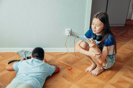 girl sitting barefoot on floor using smartphone with headphones, boy lying on floor playing on tablet smartphone, near power outlet with charging cables technology education digital lifestyleの写真素材