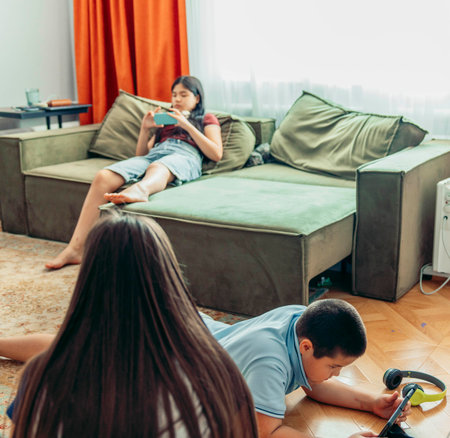 two children sitting on floor, girl lounging on sofa using digital devices, casual modern living room with orange curtains bright daylight concept of technology education leisureの写真素材