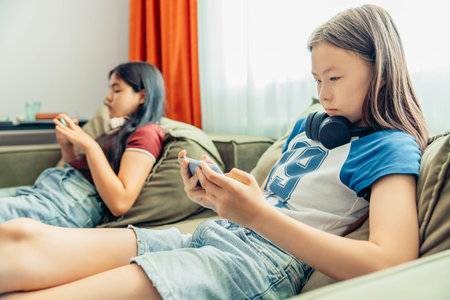 two girls relaxing on sofa using smartphone, headphones, casual home setting bright curtains and natural light window modern lifestyle, technology education entertainmentの写真素材