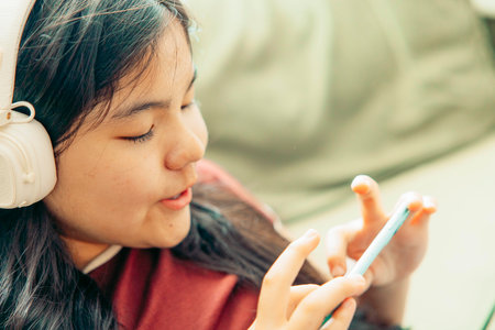 close up of Asian girl lying on green sofa using smartphone with headphones around neck focused expression home interior with soft light from window technology gadgets youth lifestyleの写真素材