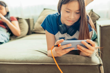 Asian half-breed girl on sofa smiling, playing on smartphone, headphones, orange charging cable connected, blurred background, sister, friend, technology education entertainmentの写真素材
