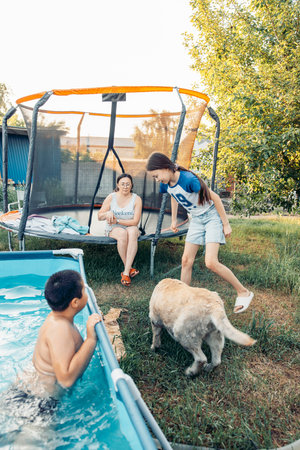 boy swimming in outdoor pool girl playing with wet labrador dog woman sitting on trampoline in sunny...