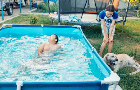 boy swimming in outdoor pool girl near pool smiling at wet labrador dog playing with plastic bottle in backyard with green grass and trampoline, family leisure pet care summer recreationの写真素材
