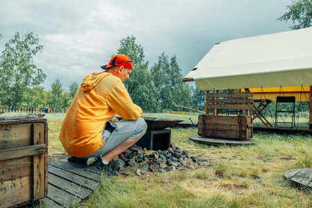 man in yellow squatting near outdoor fire pit surrounded by rocks with glamping tent in grassy field with trees, camping equipment, travel agency, outdoor adventure company, cloudsの写真素材