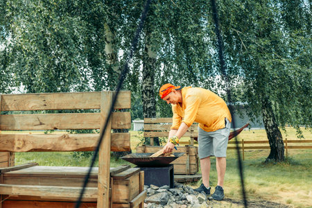 man in yellow near outdoor fire pit surrounded by stones with rustic benches seen through tent ropes, camping equipment, outdoor adventureの写真素材