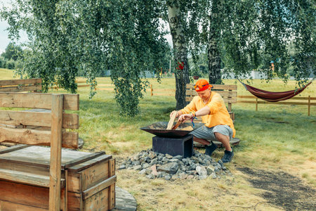 man in yellow tending outdoor fire pit with wood surrounded by rocks, rustic wooden benches in green field with birch trees and hammock, camping equipment, outdoor adventureの写真素材